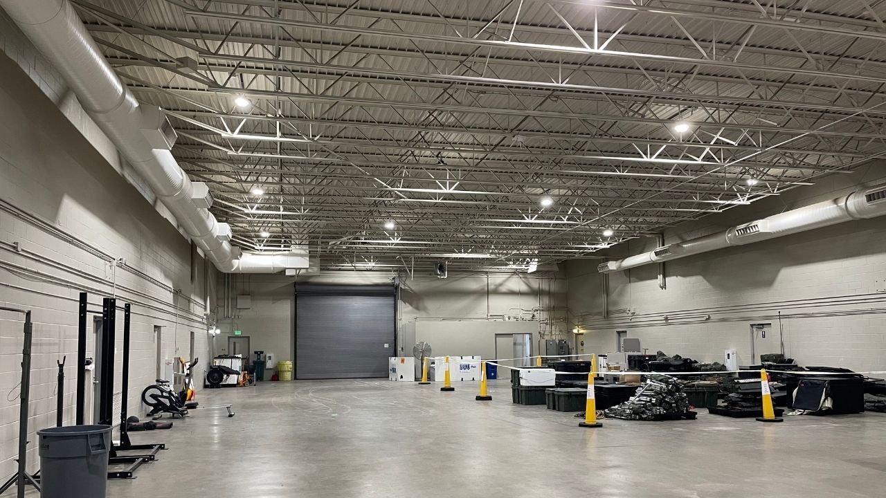 Spacious industrial interior with high ceilings, ductwork, a large garage door, and various equipment and storage items arranged along the walls and floor.