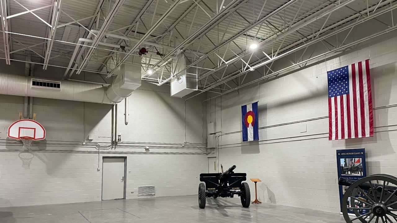 Interior of a gymnasium-like space featuring a basketball hoop, American and Colorado flags, and a historical cannon on display.